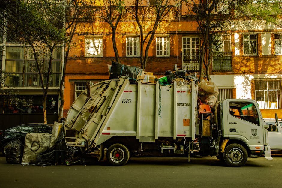 Two sanitation workers are shown loading a large blue plastic bag of waste into the rear of a refuse collection vehicle, parked alongside a city street with commercial buildings in the background. One