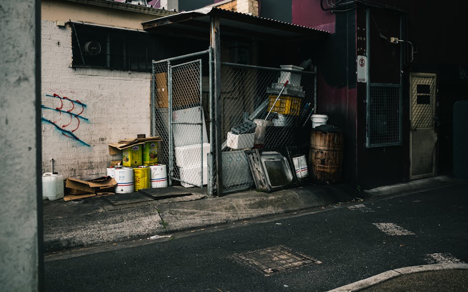 A pile of mixed waste materials is stacked against a weathered brick wall, situated in an outdoor area next to a large tree trunk with dark, textured bark. The rubbish includes flattened cardboard box
