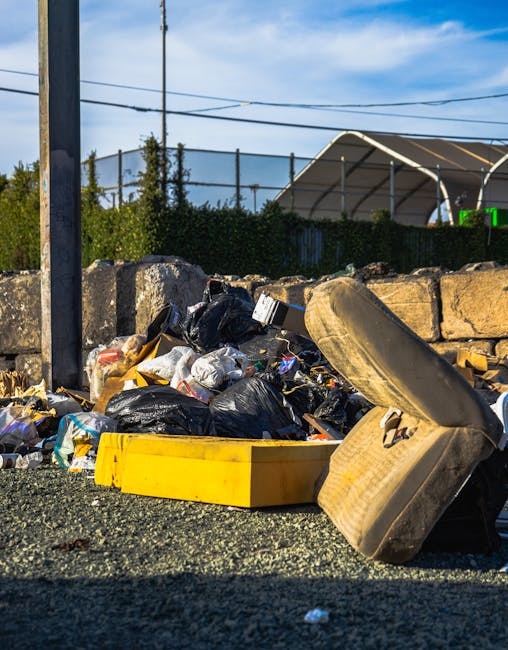 A blue flatbed truck parked on the side of a road, fully loaded with a variety of discarded household items, cardboard boxes, plastic bags, and miscellaneous waste. The truck's cab is visibly worn, wi