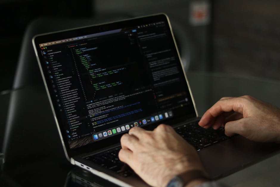 A close-up view of a person's right hand typing on a silver laptop keyboard placed on a wooden desk. The person's wrist is adorned with a patterned fabric watch strap. The laptop screen displays lines