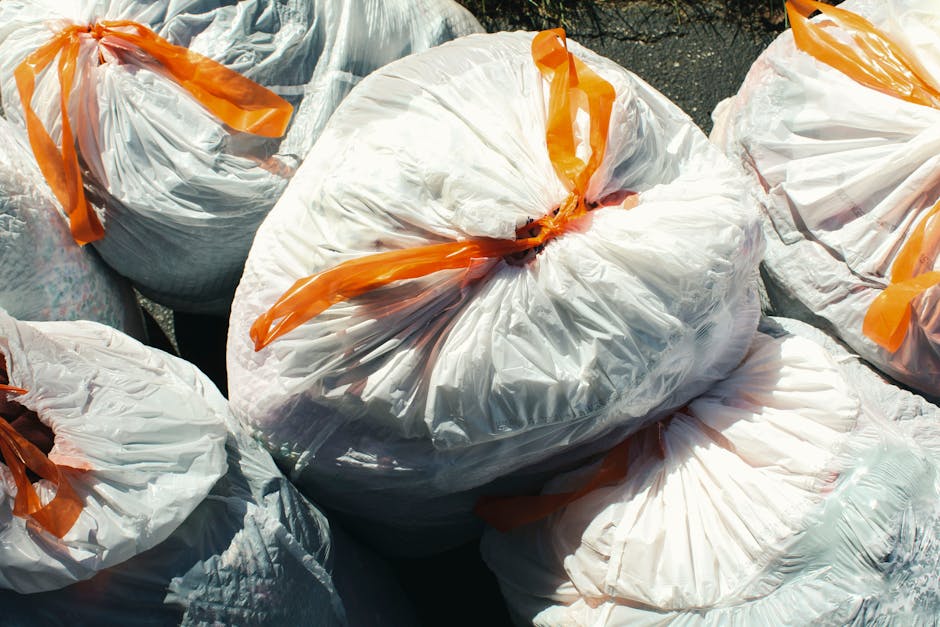 A man with dark hair and wearing a black T-shirt leans forward to empty a large white plastic bag filled with rubbish into a metal waste bin located outdoors on a paved walkway. The man is standing on